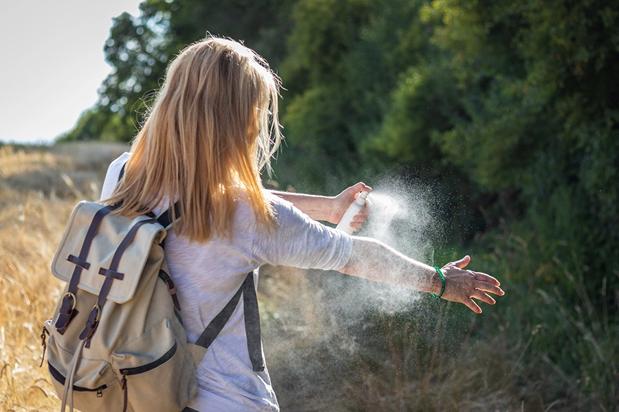A blonde woman with a beige backpack stands outdoors, spraying a white mist from a bottle onto her extended arm against a background of green trees and dry grass.