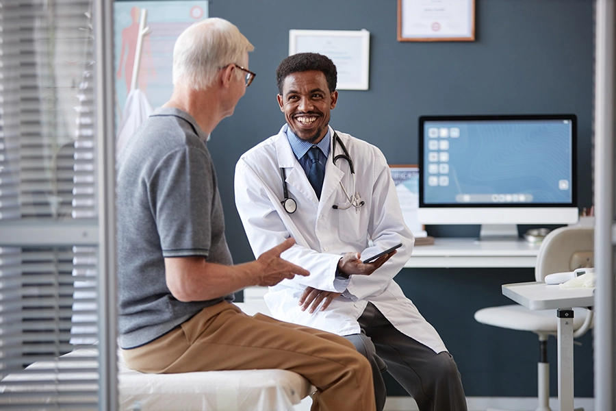 Smiling male doctor in a white coat with a stethoscope sits and talks with an older male patient during an appointment in a medical exam room.
